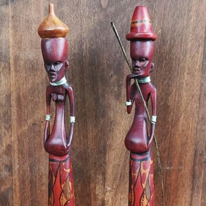 Può includere: Due statuette in legno intagliate a mano. Le figure sono marrone-rossastro con lineamenti dettagliati, che indossano collari e bracciali decorativi. Una figura tiene un bastone lungo e sottile. Le figure hanno gonne decorate in rosso, arancione e giallo.