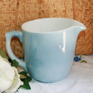 Può includere: Un'ampolla di ceramica blu chiaro con interno bianco. L'ampolla è appoggiata su un tovagliolo di pizzo bianco con un fiore bianco.