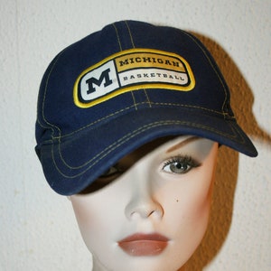 Pu&ograve; includere: Un cappello da baseball blu con una toppa gialla e bianca che dice "Michigan Basketball" e una grande "M" gialla sul davanti.