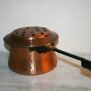 Könnte beinhalten: Ein gehämmerter Kupfertopf mit Deckel und schwarzem Griff. Der Topf hat einen runden Körper und einen breiten, flachen Boden. Der Deckel hat eine Reihe kleiner Löcher.