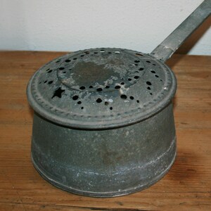 Può includere: Una pentola di metallo verde vintage con manico e coperchio forato. La pentola ha un aspetto usurato e rustico.