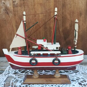 Può includere: Un modello in legno di una barca da pesca rossa e bianca con una vela bianca, salvagente neri e una base in legno. La barca è su un pizzo all'uncinetto bianco.