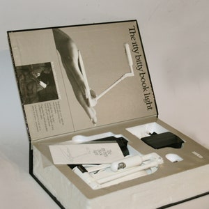 Può includere: Una luce da libro bianca nella sua confezione originale. La scatola è aperta e la luce è visibile. La scatola è etichettata "The itty bitty book light".