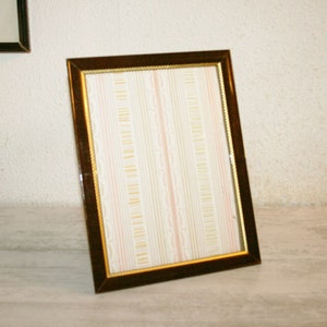 Può includere: Una cornice per foto marrone con una bordura dorata e un inserto di carta con motivo bianco e rosa.