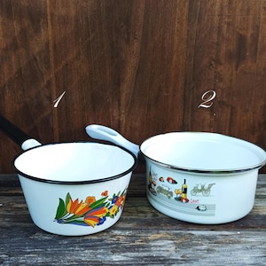 Può includere: Due pentole in smalto bianco con bordi neri e bianchi. Una pentola ha un manico nero e un motivo floreale. L'altra pentola ha un manico bianco e una fascia decorativa con immagini di auto, bottiglie e biciclette.