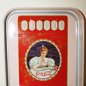 Op de afbeelding: Een roze metalen dienblad met een Coca-Cola-reclame uit 1900. De reclame toont een vrouw in een witte jurk die een glas Coca-Cola vasthoudt. Het dienblad bevat ook een kalender voor het jaar 1900. De tekst op de reclame luidt "Heerlijk en Verfrissend" en "Bij alle Soda Fountains - 5c".