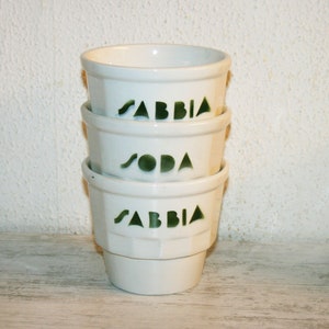 Può includere: Tre tazze in ceramica bianca impilate l'una sull'altra. Le tazze hanno un design verde con le parole "Sabbia" e "Soda" stampate su di esse.