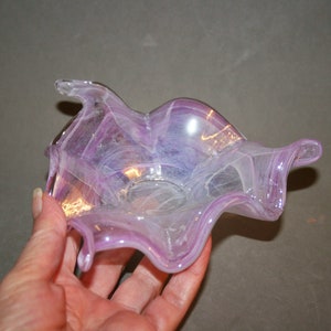 Op de afbeelding: Een kleine, handgeblazen glazen kom met een golvende, onregelmatige vorm. De kom is gemaakt van helder glas met paarse en witte strepen.