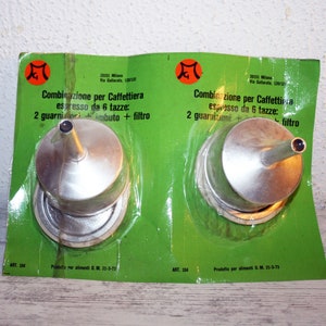 Pode incluir: Dois filtros de café de metal prateado para uma cafeteira espresso de 6 xícaras. Os filtros são embalados em um cartão verde e branco com o texto "Combinazione per Caffettiera espresso da 6 tazze: 2 guarnizioni imbuto + filtro" e "ART. 104" impresso nele.