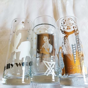 Puede incluir: Tres vasos de vidrio transparente con diseños plateados y dorados. Los vasos presentan una silueta de una mujer con un vestido, una mujer en un marco y una mujer con un rollo de película. Los vasos tienen la palabra "Hollywood" impresa en la parte inferior.