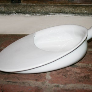 May include: A white ceramic bedpan with a smooth, glossy finish. The vessel features an elongated, oval-shaped body with a wide, flat front section that tapers into a rounded, open basin area, and includes a small, cylindrical handle at the end.
