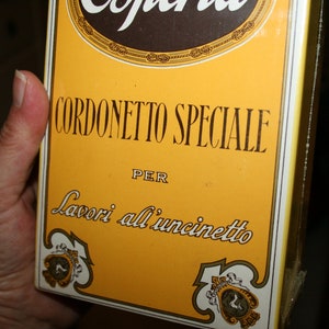 May include: A yellow box with the text "CORDONETTO SPECIALE" in black, with the word "PER" below it. The bottom of the box has a decorative emblem. The box is held by a hand.