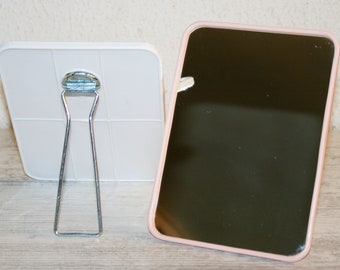 May include: Two handheld mirrors, one white and one with a pink frame. The white mirror has a metal stand.