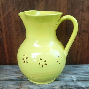 May include: A yellow ceramic pitcher with a rounded body, a curved handle, and a pouring spout. The pitcher has small, brown decorative accents and is set against a wooden background.