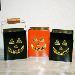Può includere: Tre portacandele di Halloween a forma di zucca. Due sono neri con facce gialle e uno è arancione con una faccia gialla. I portacandele sono in metallo e hanno un manico di filo.