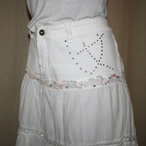 May include: A white tiered skirt with a denim waistband. The waistband has a decorative design of rhinestones in the shape of a star. The skirt has a lace trim.