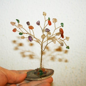 Op de afbeelding: Een draadboom sculptuur met een verscheidenheid aan kleurrijke edelstenen voor bladeren. De boom wordt getoond op een agaat plak.
