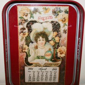 Op de afbeelding: Vintage Coca-Cola serveerblad met een rode rand en een centrale afbeelding van een vrouw die een glas Coca-Cola vasthoudt. De afbeelding is omgeven door bloemmotieven en een kalender voor april 1901. Het dienblad heeft het Coca-Cola-logo.