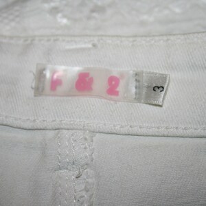 May include: White clothing label with pink lettering that reads "F&2" and a number "3" on a white background.