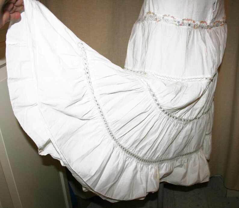 May include: A white cotton skirt with multiple layers of ruffles and lace trim. The skirt has a beaded embellishment at the waist.