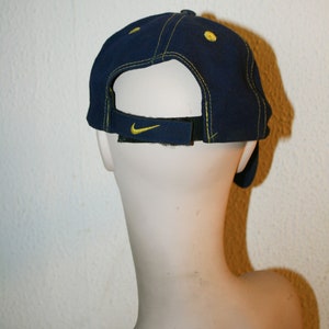 Pu&ograve; includere: Un cappello da baseball blu navy con un logo Nike Swoosh giallo sul retro. Il cappello ha un dettaglio di cucitura gialla intorno al bordo.