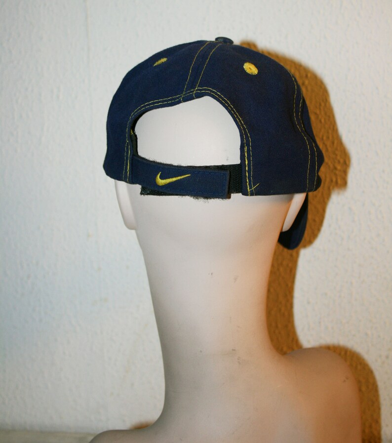 Pu&ograve; includere: Un cappello da baseball blu navy con un logo Nike Swoosh giallo sul retro. Il cappello ha un dettaglio di cucitura gialla intorno al bordo.