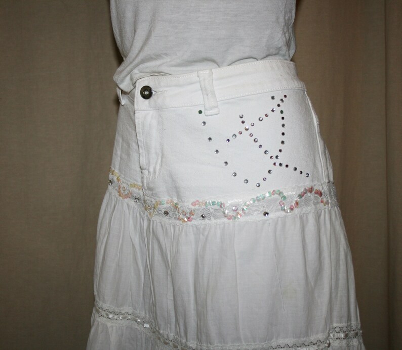 May include: A white tiered skirt with a denim waistband. The waistband has a decorative design of rhinestones in the shape of a star. The skirt has a lace trim.