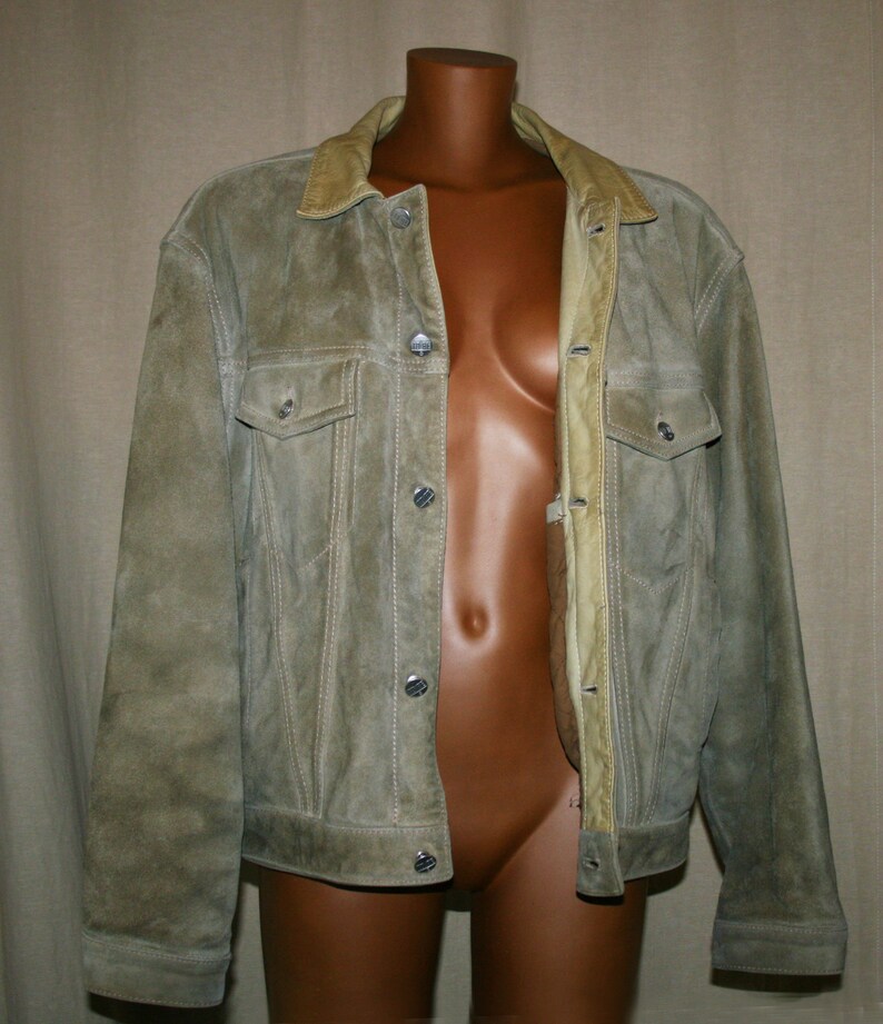 May include: A light brown suede jacket with a snap closure and a collar lined with a lighter brown suede. The jacket has two front pockets and a buttoned flap on each pocket.
