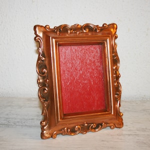May include: A copper-colored ornate picture frame with a red velvet mat. The frame has a detailed, intricate design.
