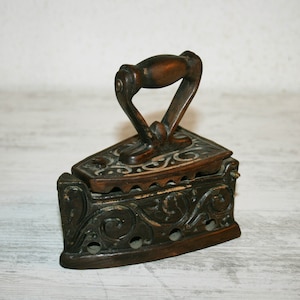 Può includere: Un ferro da stiro vintage in ghisa con un design ornamentale e un manico in legno. Il ferro è marrone e presenta un motivo dipinto in bianco.