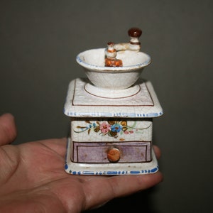 Può includere: Un macinacaffè in ceramica bianca con una manovella e un motivo floreale. Il macinacaffè ha un cassetto per riporre i chicchi di caffè.