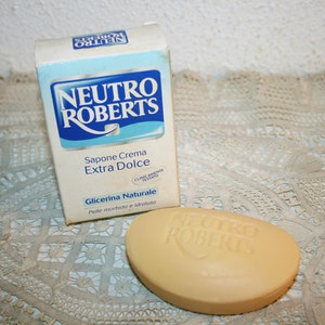 Può includere: Una scatola blu e bianca di sapone Neutro Roberts Extra Dolce con una saponetta davanti. La scatola riporta la scritta "Sapone Crema Extra Dolce" e "Glicerina Naturale". La saponetta è beige e ha il logo Neutro Roberts.