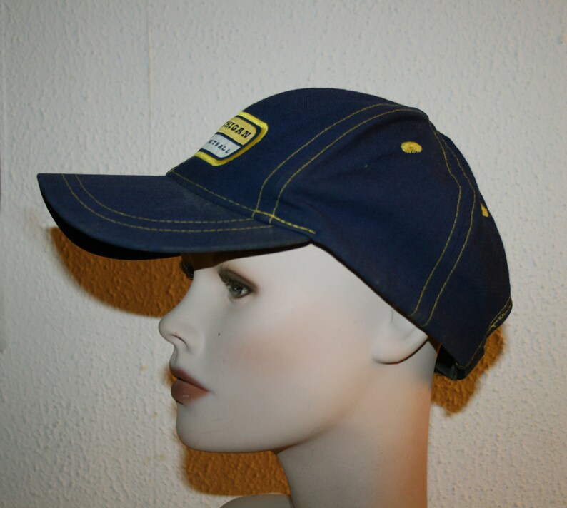 Pu&ograve; includere: Un cappello da baseball blu navy con un bordo cucito giallo e una toppa gialla che dice "Michigan Football".