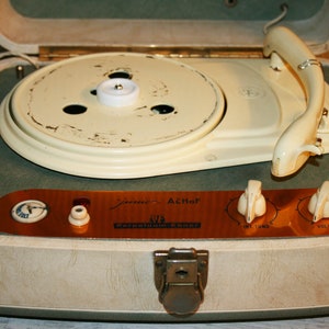 Op de afbeelding: Een vintage draagbare platenspeler in een groene en beige koffer. De speler heeft een witte draaitafel, een toonarm en een koperkleurig paneel met knoppen voor volume en toon. Het paneel heeft ook de tekst "Jannsen Althof" en "Repetierum Super" in zwart.