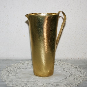 Può includere: Un'anfora dorata con una superficie testurizzata e un'unica maniglia. L'anfora è appoggiata su un pizzo bianco.