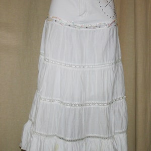 May include: White tiered skirt with a denim waistband and sequined embellishments. The skirt has a ruffled hem and is made of a lightweight fabric.