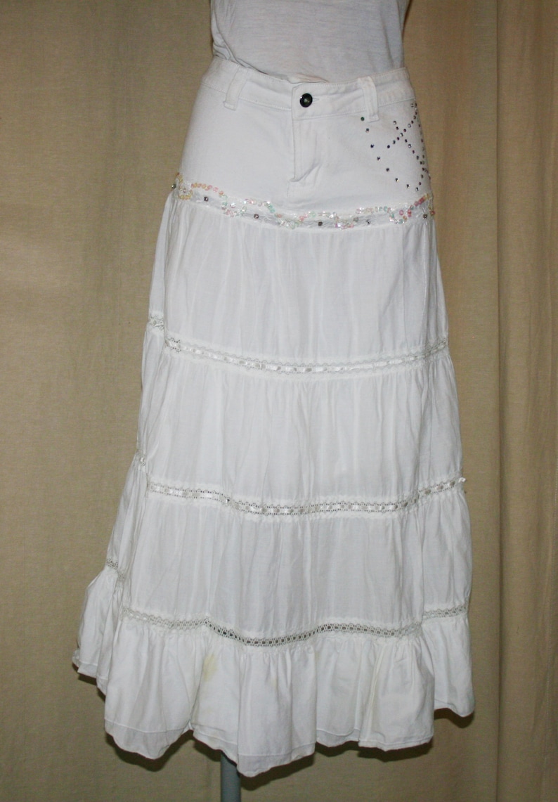 May include: White tiered skirt with a denim waistband and sequined embellishments. The skirt has a ruffled hem and is made of a lightweight fabric.