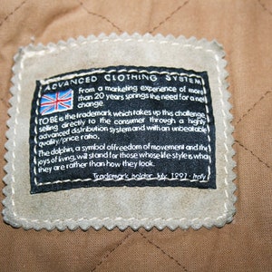 May include: A black and white label with the text "ADVANCED CLOTHING SYSTEM" and a small Union Jack flag. The label also includes the text "TO BE is the trademark which takes up this challenge selling directly to the consumer through a highly advanced distribution system and with an unbeatable quality/price ratio. The dolphin, a symbol of freedom of movement and the joys of living, we stand for those whose life style is what they are rather than how they look. Trademark holder Italy 1991 - Italy."