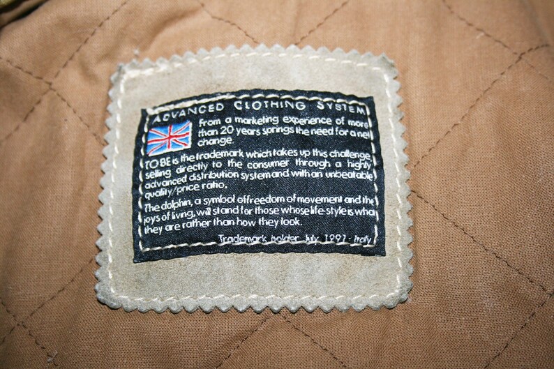 May include: A black and white label with the text "ADVANCED CLOTHING SYSTEM" and a small Union Jack flag. The label also includes the text "TO BE is the trademark which takes up this challenge selling directly to the consumer through a highly advanced distribution system and with an unbeatable quality/price ratio. The dolphin, a symbol of freedom of movement and the joys of living, we stand for those whose life style is what they are rather than how they look. Trademark holder Italy 1991 - Italy."