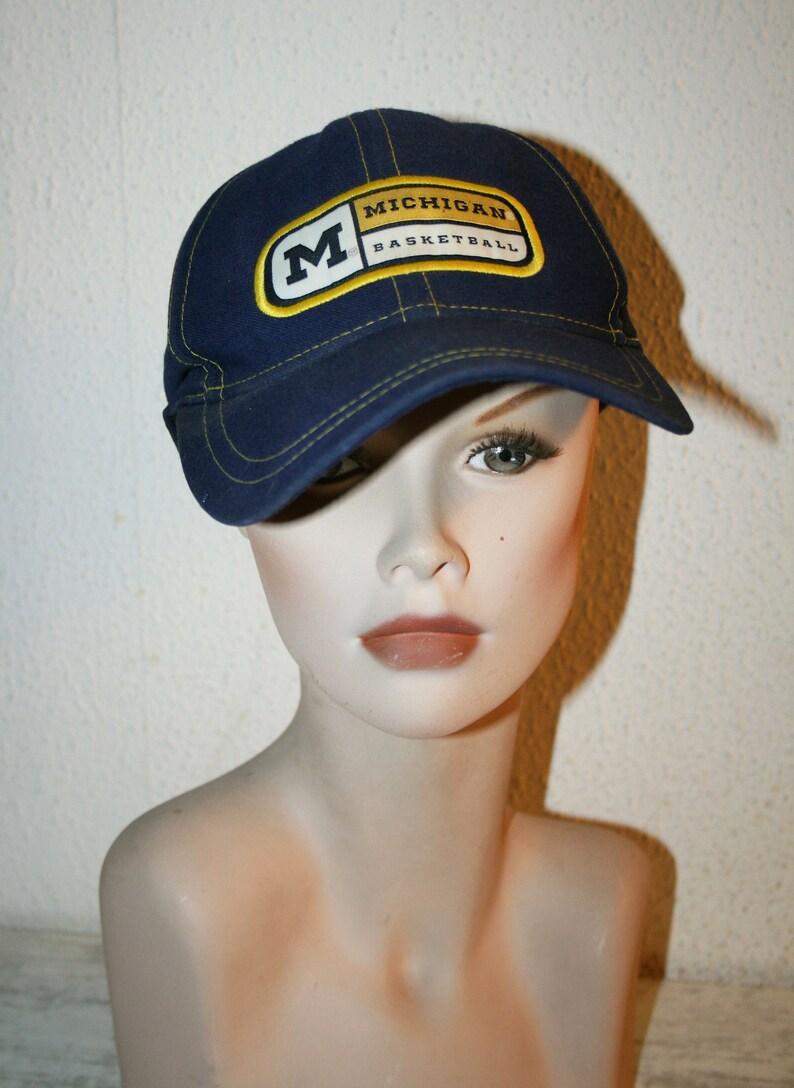 Pu&ograve; includere: Un cappello da baseball blu con una toppa gialla e bianca che dice "Michigan Basketball" e una grande "M" gialla sul davanti.