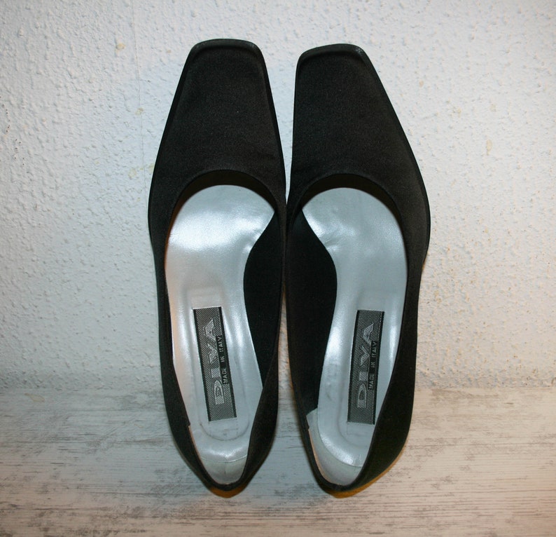 K&ouml;nnte beinhalten: Ein Paar schwarze Satin-Pumps mit spitzer Zehenpartie und niedrigem Absatz. Die Schuhe haben ein silbernes Futter und der Markenname "DIVA" ist auf der Innensohle aufgedruckt.