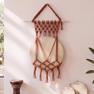 May include: A wall hanging featuring a circular wooden disc suspended by rust-colored macrame cord. The cord is knotted in a diamond pattern above and below the disc, ending in several tassels hanging from a horizontal wooden dowel rod.