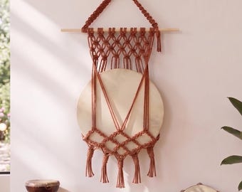 Puede incluir: Un tapiz de pared con un disco de madera circular suspendido por un cordón de macramé color óxido. El cordón está anudado en un patrón de diamante sobre y debajo del disco, terminando en varias borlas que cuelgan de una varilla de madera.