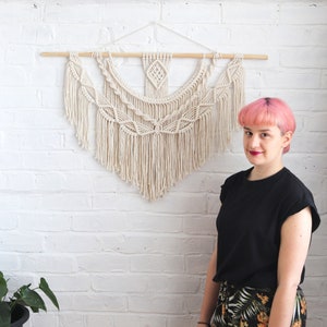 May include: A white macrame wall hanging with a geometric pattern and fringe. The hanging is mounted on a white brick wall. A person with pink hair is standing in front of the wall hanging.