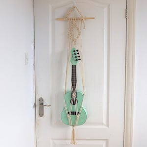 Puede incluir: Un ukelele verde menta cuelga de un colgador de macramé beige en una puerta blanca. El macramé tiene una clavija de madera en la parte superior. Un retrato en blanco y negro enmarcado cuelga en la pared a la derecha.