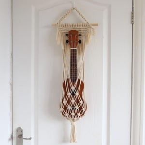 May include: A macrame ukulele holder in natural tones, suspended from a wooden dowel. The holder features a woven net design, showcasing a brown ukulele. The holder is hanging on a white door.