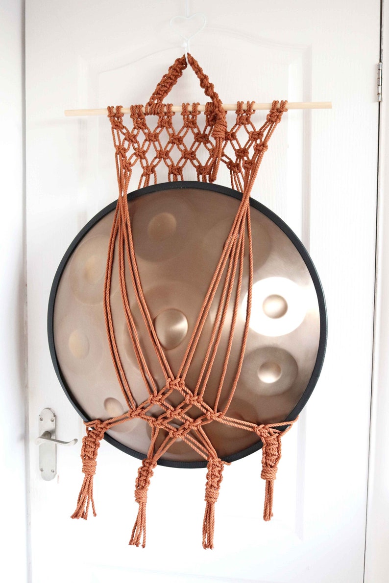 May include: A brown macrame hanging with a large, round, copper-colored handpan hanging from it. The handpan is suspended by the macrame and has a black rim.