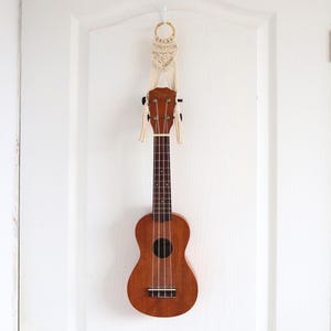 Może przedstawiać: Brązowe ukulele z ciemną podstrunnicą i czarnymi kołkami do strojenia wisi na makramie na białych drzwiach. Ukulele ma cztery struny i okrągły otwór rezonansowy. Drzwi mają białe wykończenie.