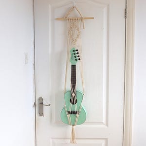 May include: A mint green ukulele hangs from a cream-colored macrame plant hanger on a white door. The ukulele has black tuning pegs and a dark brown fretboard. A framed black and white portrait hangs on the wall to the right.