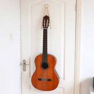 May include: A brown acoustic guitar hanging on a white door with a macrame strap. The guitar has a natural wood finish and a soundhole with a rosette.